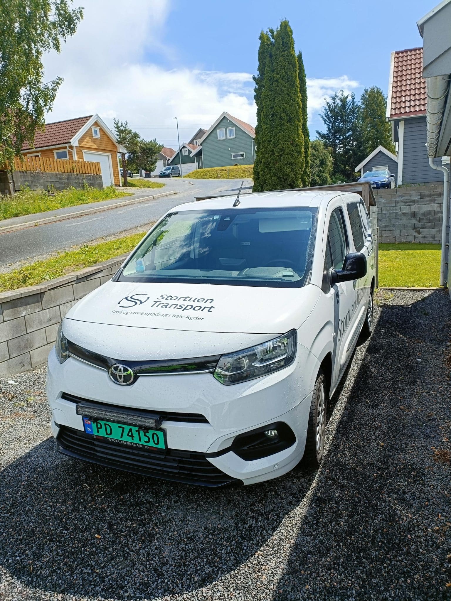 Hvit Toyota varebil med Stortuen Transport-logo parkert i boligområde, benyttet til bud- og distribusjonstjenester i Agder og omegn.